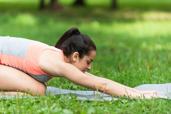 Вид збоку гнучка і підходить жінка в спортивному одязі, що тягнеться на йога мат в парку — стокове фото