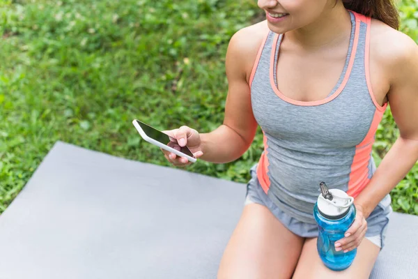 Обрізаний вид щасливої жінки, що тримає спортивну пляшку з водою і використовує смартфон у парку — Stock Photo