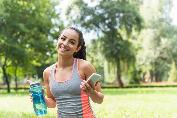 Щаслива спортсменка тримає спортивну пляшку та смартфон у парку — стокове фото