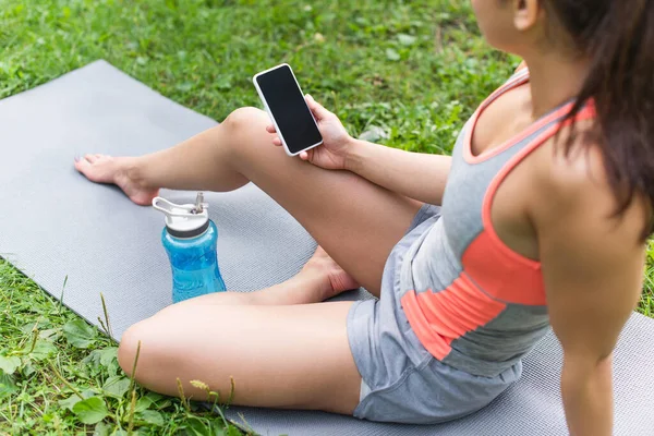 Vista recortada de la mujer usando el teléfono inteligente mientras está sentado en la esterilla de yoga cerca de la botella de deportes - foto de stock