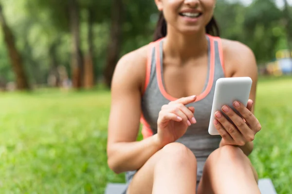 Cropped view of cheerful and blurred sportswoman using smartphone in park — Stock Photo