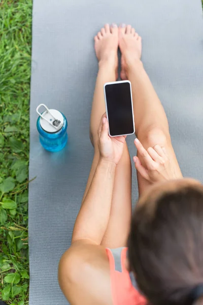 Vue du haut de la femme en utilisant un smartphone avec écran blanc tout en étant assis sur un tapis de yoga près d'une bouteille de sport — Photo de stock