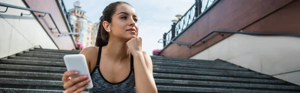 Junge Sportlerin in drahtlosen Kopfhörern mit Smartphone, während sie draußen Musik hört, Banner — Stockfoto