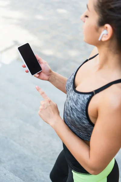 Vista de ángulo alto de la deportista en auriculares inalámbricos utilizando teléfono inteligente con pantalla en blanco fuera - foto de stock