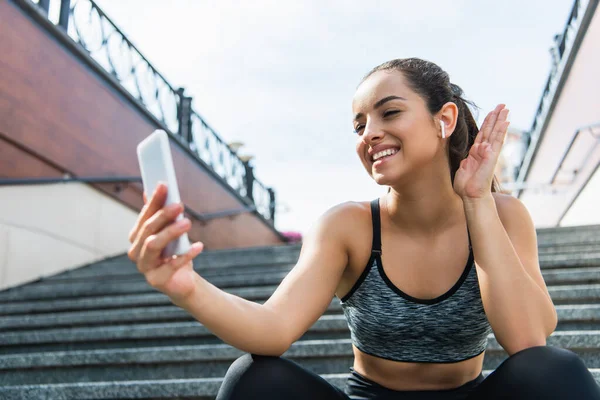 Cheerful sportswoman in wireless earphones having video call on smartphone sitting on stairs outside — Stock Photo