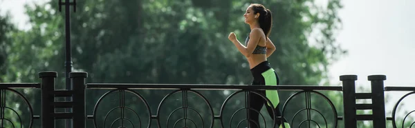 Side view of happy and sportive woman in wireless earphones listening music while running in park, banner — Stock Photo