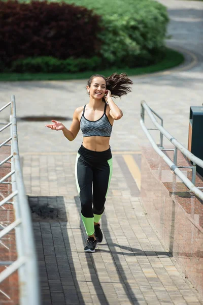 Piena lunghezza della donna felice regolazione auricolare wireless durante la corsa nel parco — Foto stock