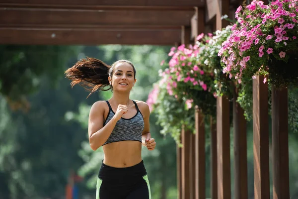 Cheerful sportswoman in wireless earphones listening music and smiling while running in park — Stock Photo