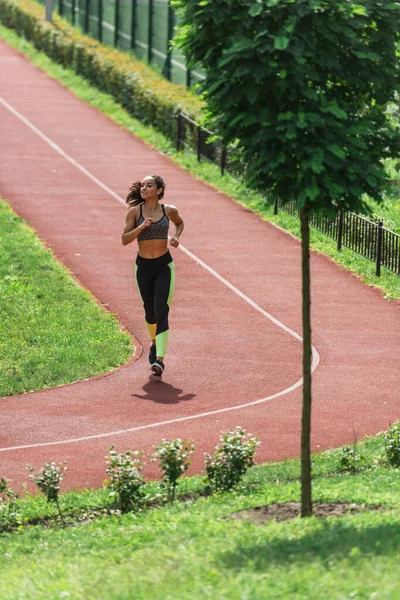 Full length of cheerful sportswoman in wireless earphones listening music while running on stadium — Stock Photo