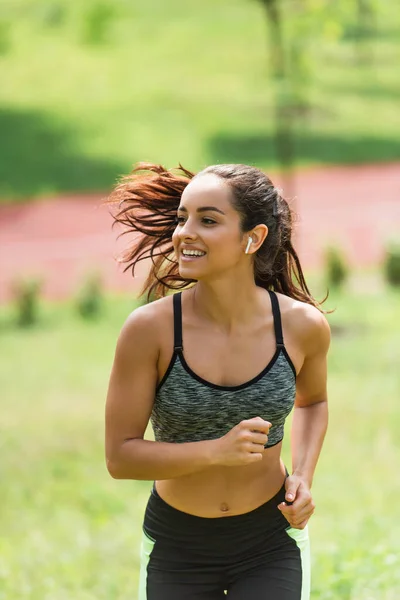 Joyful sportswoman in crop top and wireless earphones running in park — Stock Photo