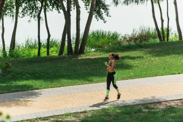 Full length of happy sportswoman in wireless earphones listening music while running in green park — Stock Photo