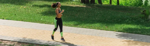 Piena lunghezza di donna sportiva felice in auricolari wireless in esecuzione nel parco verde, banner — Foto stock
