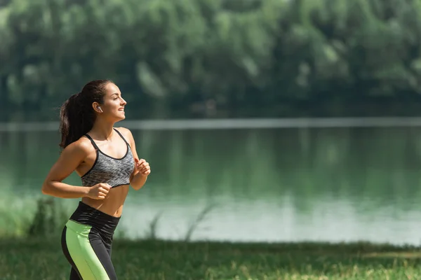 Vue latérale de la sportive heureuse dans les écouteurs sans fil écouter de la musique tout en courant dans un parc verdoyant près du lac — Photo de stock