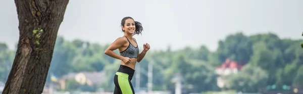 Fröhliche Sportlerin in bauchfreiem Top und Leggings beim Joggen im Park, Banner — Stockfoto