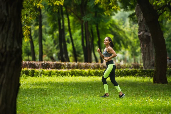 Volle Länge der fröhlichen Sportlerin in Crop Top und Leggings beim Joggen auf dem Rasen im Park — Stockfoto