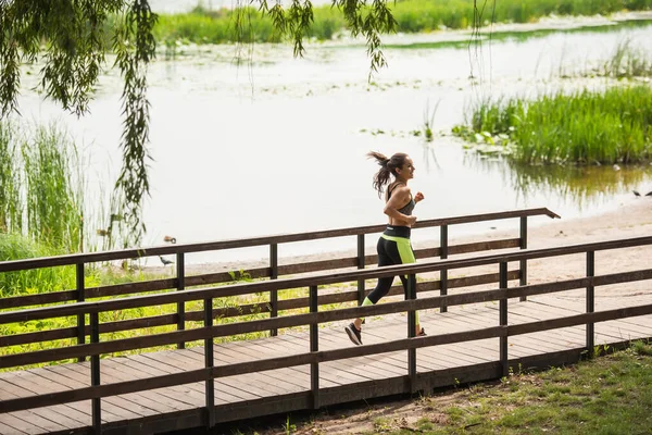 Longitud completa de la mujer feliz en la parte superior de la cosecha y leggings trotar en el puente cerca del lago en el parque - foto de stock