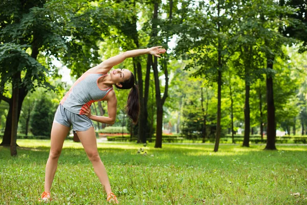 Piena lunghezza di donna felice e sportiva che si esercita con mano sull'anca mentre si riscalda nel parco — Foto stock