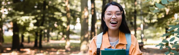 Aufgeregte asiatische Frau mit Notizbüchern, die im Park vor laufender Kamera lacht, Banner — Stockfoto