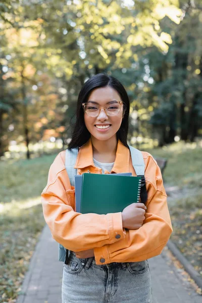 Brunette asian student in eyeglasses smiling at camera while holding notebooks in park — Stock Photo