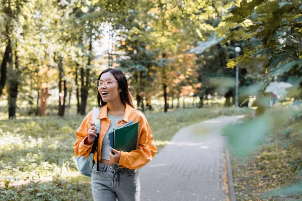 Stupéfait asiatique étudiant dans lunettes regarder loin tout en marchant dans parc avec copie livres — Photo de stock