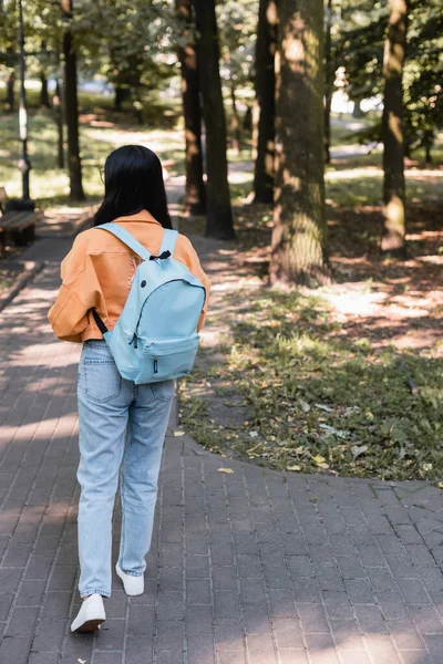 Vista posteriore dello studente in jeans che cammina nel parco con zaino — Foto stock