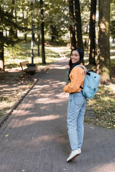 Piena lunghezza vista di asiatico studente in arancione giacca e jeans sorridente a fotocamera in parco — Foto stock