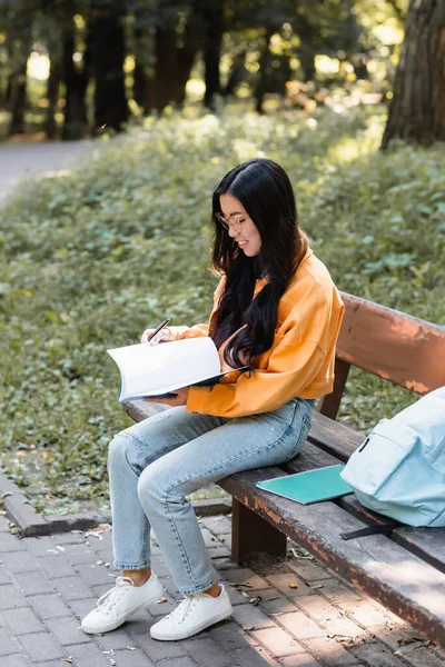 Smiling asian student sitting with notebook near backpack on bench in park — Stock Photo