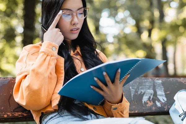 Thoughtful asian student adjusting eyeglasses while looking in notebook in park — Stock Photo