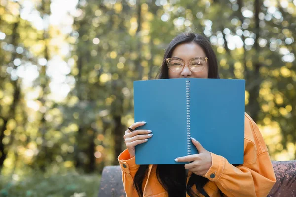 Cheerful asian student in eyeglasses obscuring face with copy book in park — Stock Photo
