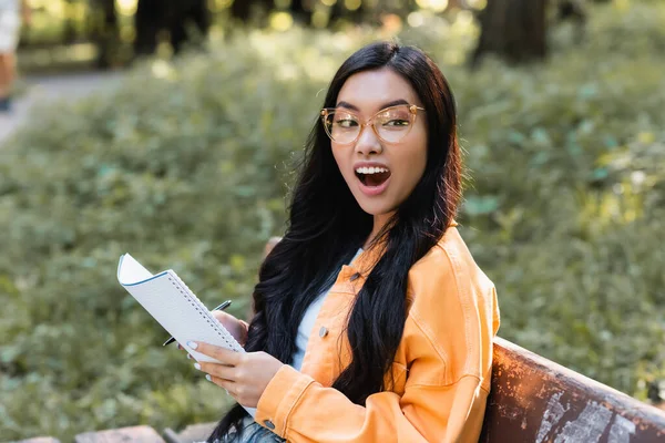 Asombrado asiático estudiante con notebook mirando lejos mientras sentado en banco en parque - foto de stock