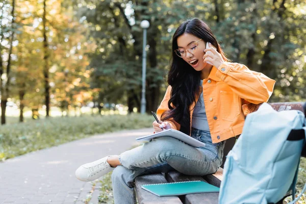 Asombrada mujer asiática escribiendo en el cuaderno mientras habla en el teléfono inteligente en el banco en el parque - foto de stock