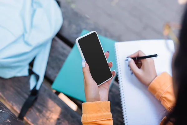 Teilansicht des Schülers, der in Notizbuch schreibt, während er sein Smartphone mit leerem Bildschirm im Freien hält — Stockfoto
