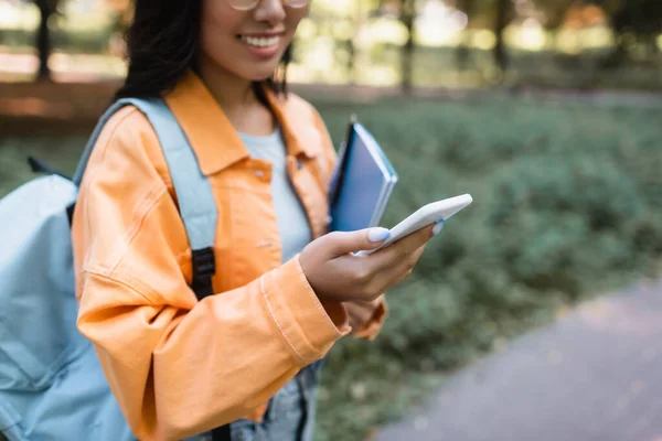 Visão parcial da mulher desfocada em casaco laranja usando telefone celular ao ar livre — Fotografia de Stock