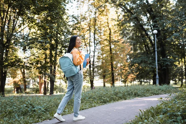 Улыбающаяся азиатка в джинсах, гуляющая в парке с рюкзаком и блокнотами — стоковое фото