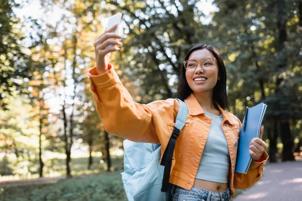 Cheerful asian woman with backpack and notebooks taking selfie on cellphone in park — Stock Photo