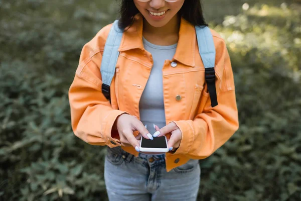 Vista recortada de la mujer sonriente en chaqueta naranja utilizando el teléfono celular con pantalla en blanco - foto de stock