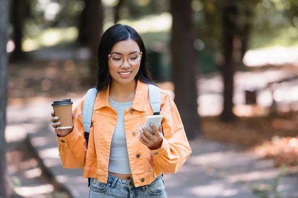 Felice donna asiatica guardando il telefono cellulare mentre tiene take away bere all'aperto — Foto stock