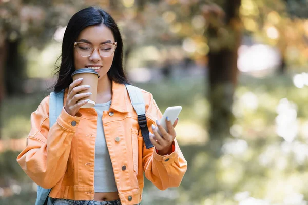 Positive asian student drinking coffee to go while messaging on cellphone in park — Stock Photo