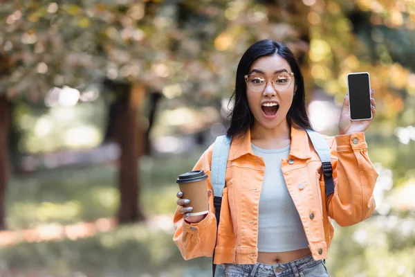 Stupito asiatico studente con caffè a andare mostrando cellulare con vuoto schermo in parco — Foto stock