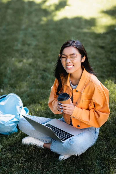 Alegre asiático mujer en gafas sentado en hierba con portátil y café para ir - foto de stock