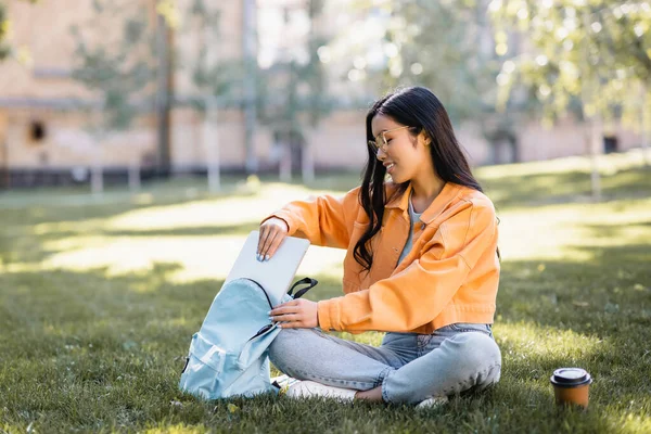 Souriant asiatique femme mettre ordinateur portable dans sac à dos tout en étant assis sur l'herbe avec les jambes croisées — Photo de stock