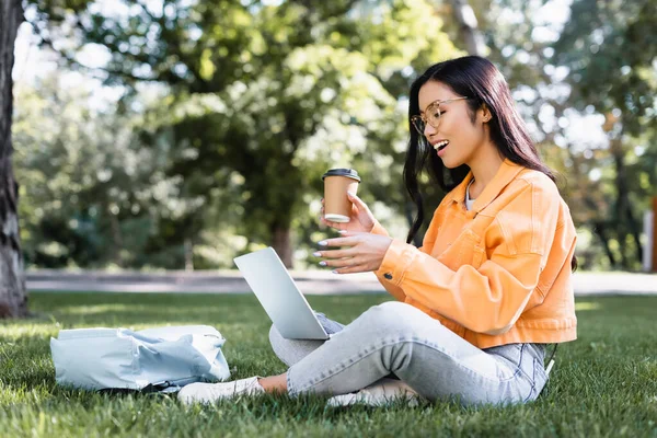 Excited asian student pointing at laptop while sitting on lawn with coffee to go — Stock Photo