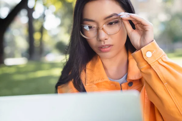 Pensativo asiático mujer tocando gafas mientras mirando borrosa portátil al aire libre - foto de stock