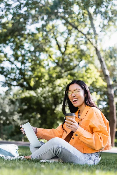 Excited asian student with takeaway drink laughing while sitting on lawn with laptop — Stock Photo