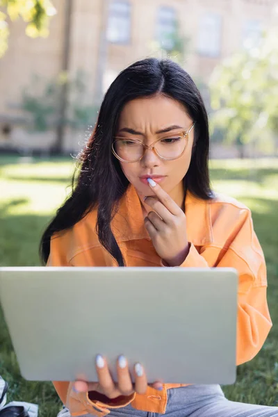 Reflexivo asiático mujer en gafas tocando labios mientras mira portátil en parque - foto de stock