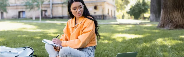 Sonriente asiático estudiante escritura en notebook cerca de portátil y mochila en hierba, bandera - foto de stock