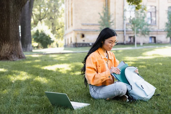 Молодая азиатка сидит на траве возле ноутбука и кладет тетради в рюкзак — стоковое фото