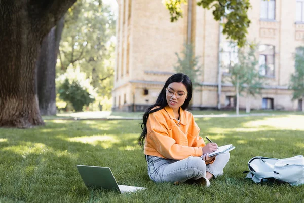 Asiatin schreibt in Notizbuch, während sie auf Rasen in der Nähe von Laptop und Rucksack sitzt — Stockfoto