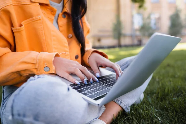 Частичный вид студента в оранжевой куртке, сидящего в парке и печатающего на ноутбуке — стоковое фото