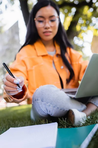 Blurred asian woman sitting on lawn with laptop and pen near notebooks — Stock Photo